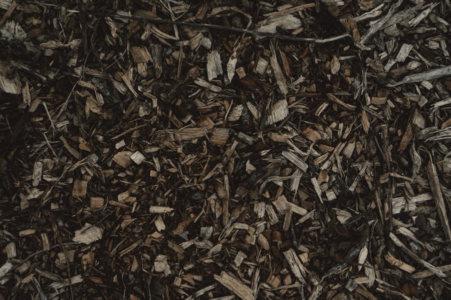 A person uses a metal garden rake to lightly turn and aerate dark brown wood chip mulch in a landscaped garden bed.