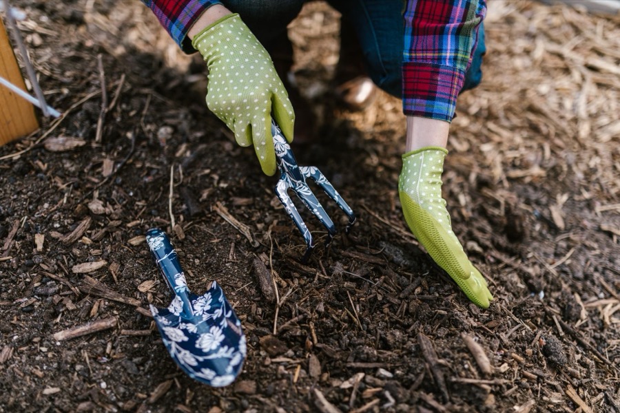 A person using a garden rake to gently aerate and turn over dark brown wood mulch in a garden bed.